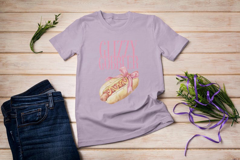 Puede incluir: Una camiseta lavanda con el texto "Gizzy Gourmet" en letras rosas. Una caricatura de un hot dog con mostaza y un lazo rosa est&aacute; impresa en la camiseta.