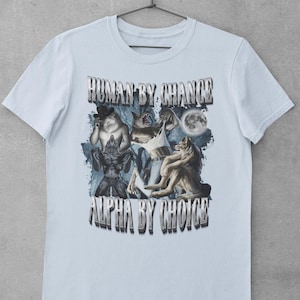 May include: A light blue short-sleeve t-shirt featuring a graphic print of multiple wolves and a full moon. The text "HUMAN BY CHANCE" is printed above the graphic, and the text "ALPHA BY CHOICE" is printed below the graphic in a metallic font.