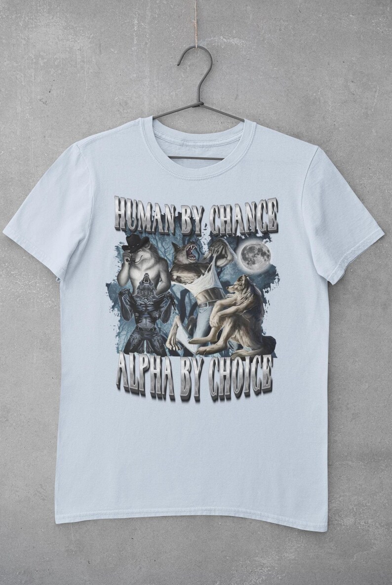 May include: A light blue short-sleeve t-shirt featuring a graphic print of multiple wolves and a full moon. The text "HUMAN BY CHANCE" is printed above the graphic, and the text "ALPHA BY CHOICE" is printed below the graphic in a metallic font.