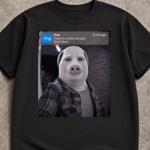 Puede incluir: Una camiseta negra con un estampado gráfico de una notificación de timbre Ring. El estampado muestra una imagen en blanco y negro de una persona con cara de cerdo y camisa de cuadros, con el texto Ring, There is motion at your Front Door, 8 min ago.
