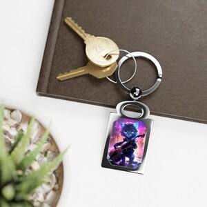 Puede incluir: Un llavero de plata con un dije rectangular que presenta un gato de dibujos animados tocando un violonchelo en un entorno urbano. El gato es azul con ojos verdes y lleva una camisa roja. El fondo es un paisaje urbano morado y azul.