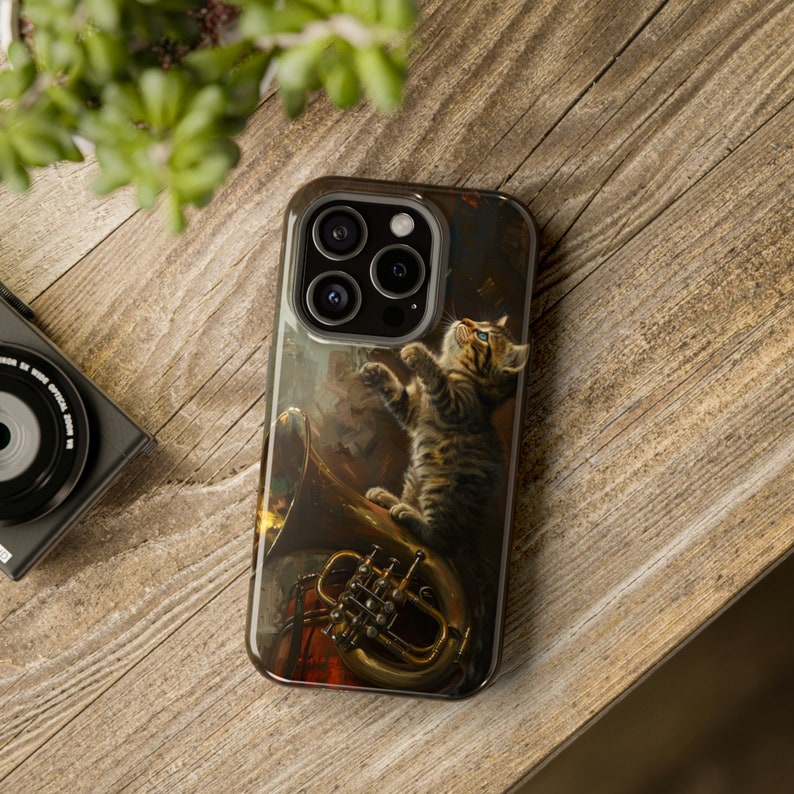 May include: A phone case with a colorful illustration of a cat playing a musical instrument. The cat is brown and white with green eyes. The instrument is a gold-colored French horn. The case has a black background and a camera lens cutout.