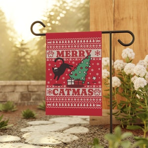 May include: A red Christmas garden flag featuring a black cat wearing a Santa hat knocking over a green Christmas tree. The flag has a white background with a red and white snowflake pattern and the text "Merry Catmas".