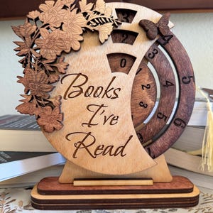 May include: A circular wooden book tracker with the text Books I've Read engraved on the front. The design features a laser-cut floral arrangement on the left, a butterfly on the right, and rotating numbered dials to track the number of books read.