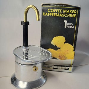 May include: A silver and black one-cup coffee maker with a gold-coloured spout. The box behind it reads "COFFEE MAKER KAFFEEMASCHINE" and features a yellow flower illustration. The coffee maker is a vintage style.