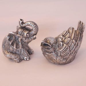 Op de afbeelding: Twee zilverkleurige metalen beeldjes, bestaande uit een zittende olifant met de slurf omhoog en een vogel met gedetailleerde, gelaagde vleugelveren. Beide objecten hebben een textuurrijke, metalen afwerking met zichtbare lijnen.