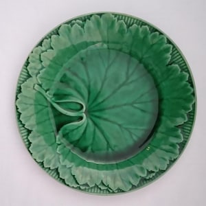 Puede incluir: Un plato redondo de cerámica con un esmalte verde vibrante y un diseño en relieve detallado de hojas superpuestas. El centro muestra una hoja prominente con venas visibles, rodeada por un borde festoneado con un patrón de rejilla.