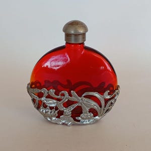 Puede incluir: Una botella redonda de vidrio rojo translúcido con un revestimiento de metal plateado. La carcasa decorativa de metal muestra intrincados motivos florales y de libélulas que envuelven la parte inferior, rematada con un tapón plateado.