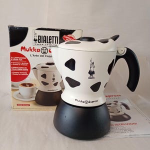 Pode incluir: Uma cafeteira de cappuccino Bialetti Mukka Express com corpo superior branco com manchas pretas de vaca, base preta e alça preta. O produto é exibido ao lado da embalagem original de papelão e de um manual de instruções.