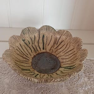May include: A ceramic bowl shaped like a flower with scalloped edges. The interior features a dark brown, textured center surrounded by light tan petals with deep, vertical grooves and subtle green accents radiating toward the rim.