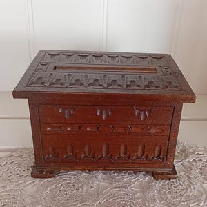 May include: A rectangular dark brown wooden box with a coin slot on the top lid. The exterior features carved geometric patterns along the lid edges and horizontal bands of repeating decorative motifs across the front and top surfaces.