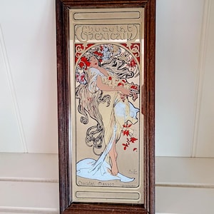 Peut inclure: Un miroir mural rectangulaire dans un cadre en bois foncé présentant une impression décorative d'une personne aux longs cheveux flottants entourée d'éléments floraux rouges et blancs. Le texte Chocolat Mexicain et Chocolat Masson est imprimé.