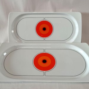 Könnte beinhalten: Zwei rechteckige weiße Servierplatten aus Keramik mit abgerundeten Ecken. Jede Platte zeigt ein zentrales grafisches Design, bestehend aus einem schwarzen Punkt, umgeben von konzentrischen orangefarbenen und roten Kreisen, eingerahmt von einer dünnen schwarzen Linie.
