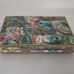 Op de afbeelding: Een rechthoekige doos met een mozaïek van iriserende abalone schelpstukjes op het deksel en de zijkanten. De schelpfragmenten vertonen een verscheidenheid aan kleuren, waaronder groen, blauw, roze en paars, gevat in een dun, zilverkleurig frame.