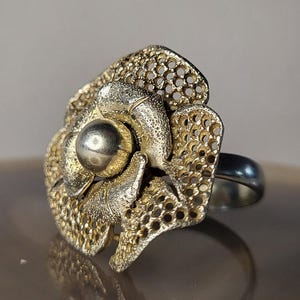 Puede incluir: Un anillo de tono dorado con una pieza central grande y dimensional en forma de flor. Los pétalos tienen un diseño calado texturizado tipo panal con una cuenta esférica lisa y pulida en el centro. La banda es de metal liso y pulido.