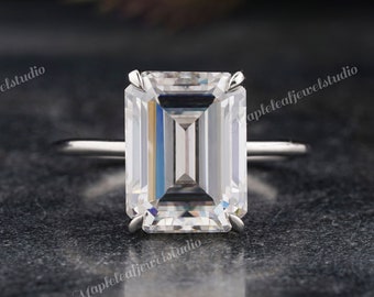 May include: A silver engagement ring featuring a large, rectangular emerald-cut diamond. The clear diamond reflects light, set on a thin, polished band. The ring is displayed against a dark, textured background.