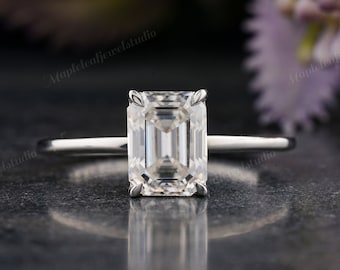 May include: A solitaire engagement ring with an emerald-cut diamond. The diamond is set on a slender, polished silver band. The ring is shown on a dark, textured surface with blurred floral accents in the background.