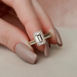 May include: A gold engagement ring with a large, rectangular, clear gemstone. A smaller band with small, clear gemstones sits next to the engagement ring. The rings are held up against a soft, neutral background.