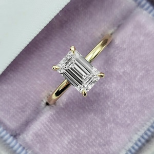 May include: A gold ring with an emerald-cut diamond. The diamond is clear and rectangular, set with four prongs. The ring is in a purple velvet box.