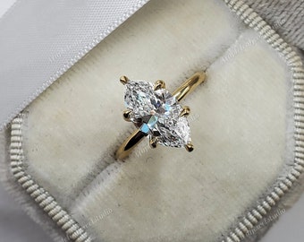 May include: A gold engagement ring featuring two marquise-cut diamonds. The ring is presented in a grey velvet box, with a white ribbon. The diamonds are cut to reflect light, creating a sparkling effect.