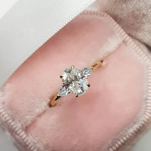 Könnte beinhalten: Ein goldener Verlobungsring mit einem großen, oval geschliffenen Diamanten und zwei birnenförmigen Seitensteinen. Der Ring ist in einem zarten Goldband gefasst und liegt in einer rosa Samt-Ringbox.