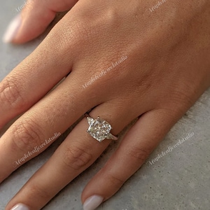 Puede incluir: Anillo de compromiso de plata con un gran diamante central de talla cuadrada y dos diamantes triangulares más pequeños a los lados. El anillo se muestra en una mano con uñas cuidadas.