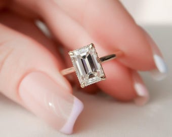 May include: A gold ring featuring an emerald-cut diamond. The clear, rectangular diamond is set in a gold band. The ring is held by a person with manicured nails. The ring is a piece of jewellery.