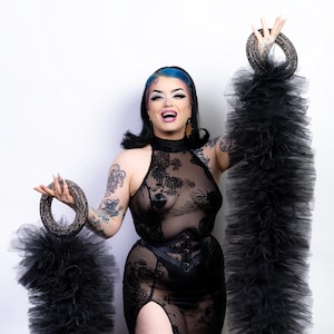 Könnte beinhalten: Eine Person in einem schwarzen, durchsichtigen Kleid mit floralem Muster, hohem Schlitz und Korsett. Sie hält zwei lange, schwarze, gerüschte Accessoires. Die Person hat Tätowierungen, blaue Haare und trägt Make-up.