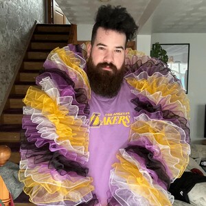 May include: A person wearing a purple shirt with the text "Los Angeles Lakers" on the front. The person is wearing a large, multi-layered ruffle collar in yellow, white, purple, and black.