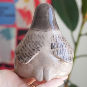 May include: A ceramic bird figurine in shades of gray and brown. The bird has a rounded body and a smooth, glossy finish. The wings are detailed with a textured pattern. The figurine is held in a person's hand.