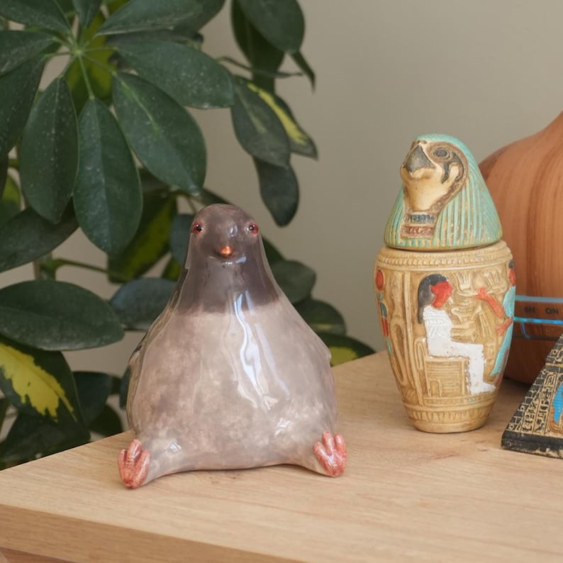May include: A ceramic pigeon figurine with a gray body and a dark gray head. The bird has red eyes and feet, and is sitting down. An Egyptian-style jar and a pyramid are in the background.
