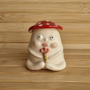 May include: A white ceramic figurine of a mushroom person with a red and white spotted mushroom hat. The figurine is holding a small bouquet of pink flowers.