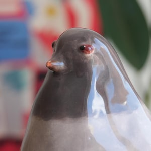 May include: A ceramic bird figurine with a glossy finish. The bird has a dark gray head and a lighter gray body. It features red eyes and a small, orange beak. The figurine is a decorative item.