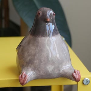 May include: A ceramic pigeon figurine with a glossy finish. The bird is primarily gray with a darker gray head and red feet and beak. The pigeon is sitting with its legs extended, giving it a whimsical appearance.
