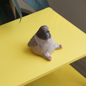 May include: A ceramic pigeon figurine with a gray body, dark gray head, and red feet and eyes. The bird is sitting on a bright yellow surface. The figurine has a whimsical, cartoonish design.