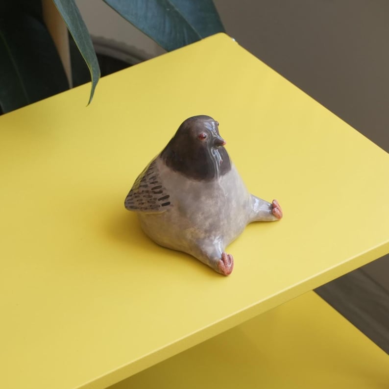 May include: A ceramic pigeon figurine with a gray body, dark gray head, and red feet and eyes. The bird is sitting on a bright yellow surface. The figurine has a whimsical, cartoonish design.