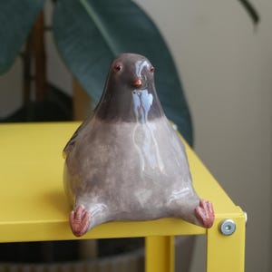 May include: A ceramic pigeon figurine with a glossy finish. The bird is primarily gray with a darker gray head and a small orange beak. Its feet are a reddish-brown color. The pigeon is sitting on a yellow surface.