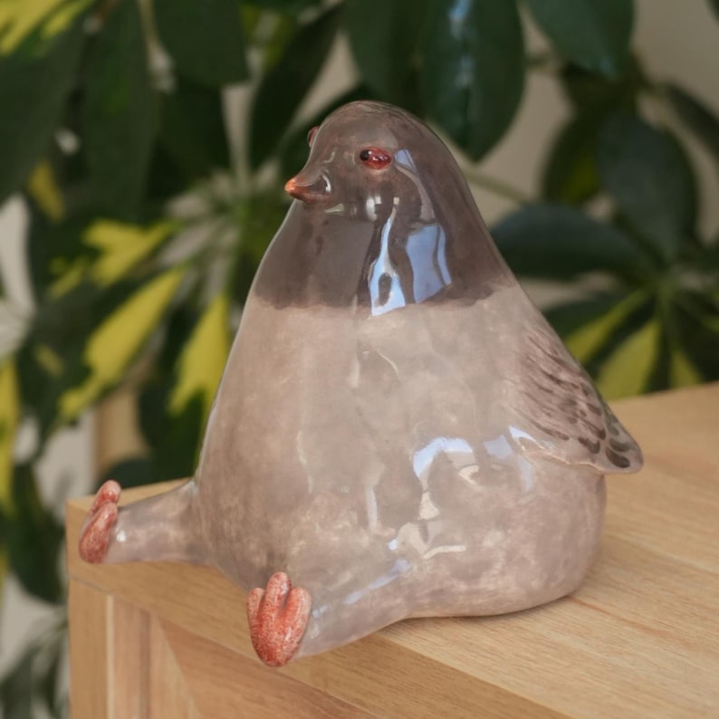 May include: A ceramic bird figurine with a rounded body, featuring a two-tone color scheme of light gray and dark brown. The bird has small, orange feet and a matching beak. The figurine is sitting on a wooden surface.