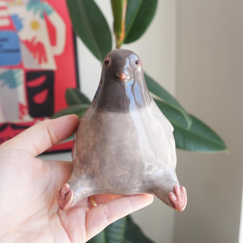 May include: A ceramic pigeon figurine with a gray body and a darker gray head. The bird has small, reddish-brown feet and a glossy finish. The figurine is held in a hand, showcasing its unique design and craftsmanship.