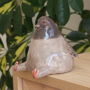 May include: A ceramic bird figurine with a rounded body, gray and brown coloring, and small red feet. The bird is perched on a wooden surface, with green foliage in the background. The figurine has a glossy finish.