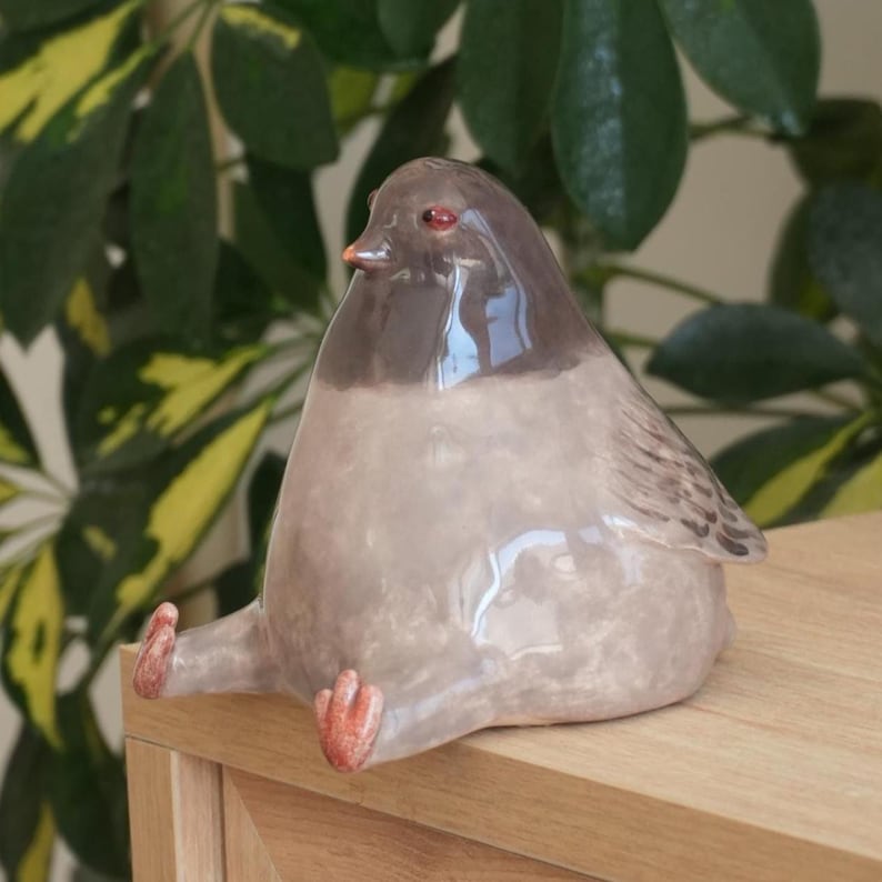 May include: A ceramic bird figurine with a rounded body, gray and brown coloring, and small red feet. The bird is perched on a wooden surface, with green foliage in the background. The figurine has a glossy finish.