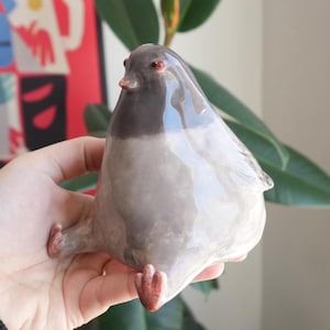 May include: A ceramic pigeon figurine in shades of gray, with a dark gray head and red eyes. The bird is sitting with its legs extended. The figurine has a glossy finish and is a decorative item.
