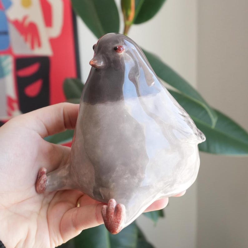May include: A ceramic pigeon figurine in shades of gray, with a dark gray head and red eyes. The bird is sitting with its legs extended. The figurine has a glossy finish and is a decorative item.