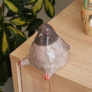 May include: A ceramic pigeon figurine with a gray body and a dark gray head. The bird has red eyes and small, orange feet. The figurine is sitting on a wooden surface.