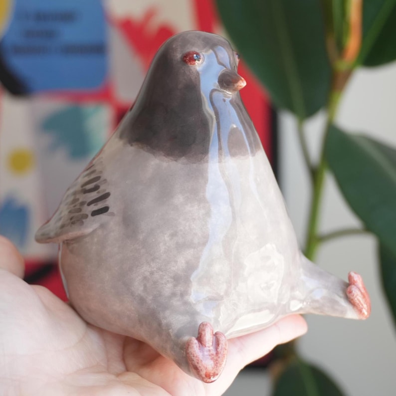 May include: A ceramic pigeon figurine with a glossy finish. The bird is primarily gray with a dark gray head and wings. It has red eyes and pink feet. The figurine is sitting with its feet extended.
