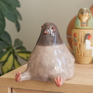 May include: A ceramic pigeon figurine with a gray body and a dark gray head. The bird has red eyes and feet, and is sitting with its legs extended. The figurine is on a wooden surface.