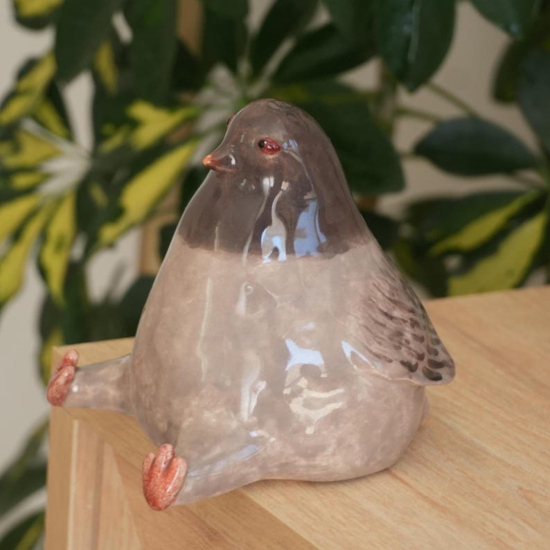 May include: A ceramic bird figurine with a glossy finish. The bird is primarily light gray with a dark brown head and wings. It has small, orange feet and a beak, and is sitting down. The figurine is on a wooden surface.