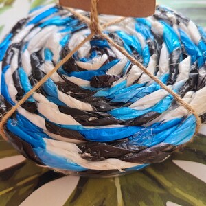 Puede incluir: Un posavasos redondo hecho de tiras entrelazadas de plástico azul, blanco y negro. El posavasos está atado con hilo y tiene una pequeña etiqueta marrón. Diseñado para proteger las superficies del calor y la humedad.