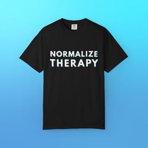 Può includere: T-shirt nero con la scritta bianca "NORMALIZE THERAPY". La maglietta a maniche corte ha lo scollo tondo. Lo sfondo è un gradiente blu.