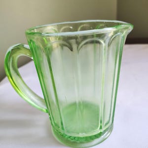 May include: A vintage green glass creamer with a handle. The creamer has a ribbed design and a rounded base. The glass is a light, translucent green colour. The creamer is likely from the Art Deco era.